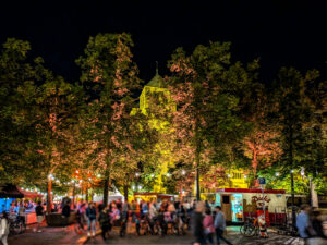 Blick von Süd Südost über den Domplatz auf den Dom von Münster. Es ist später Abend und der Himmel ist dunkel. Der Dom und die Bäume am Domplatz sind farbig illuminiert. Im Vordergrund sind einige gastronomische Stände zu sehen, es ist sehr belebt.