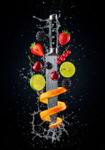 Ein wertvoll aussehendes Küchenmesser durchstößt mit der unteren Hälfte seiner Klinge senkrecht von oben nach unten eine spiralförmig aufgeschnittene Orange. Die obere Hälfte der Klinge und der Griff sind umgeben von weiteren Früchten wie Kirschen, Johannisbeeren, Brombeeren, Erdbeeren sowie Limettenscheiben. Außerdem spritzen Wasser und Wassertropfen in alle Richtungen. Der Hintergrund ist schwarz.