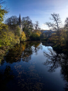 Blick auf die Ems in Telgte. Links der Ems ist der Turm der St. Clemens-Kirche zu sehen. Die Bäume und Sträucher am Rand der Ems sind herbstlich gefärbt oder schon ohne Laub und spiegeln sich in dem ruhigen Wasser der Ems.