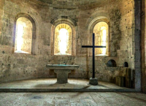Blick in die Apsis der Abteikirche der Abbaye du Thoronet. Zu sehen sind in dem Halbrund vor drei Fenstern im romanischen Stil ein schlichter Altar, rechts davon stehend ein großes Kreuz.