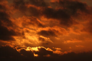 Das Bild zeigt einen wolkenreichen Himmel. Die Wolken und der ganze Himmel erscheinen durch die hinter den Wolken untergehende Sonne von Gelb über Orange, Braun-Rot bis zu Schwarz in verschiedenen Farbtönen.