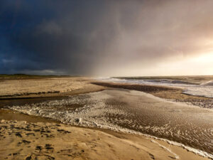 Blick auf die Grenzlinie zwischen Meer und Strand. Links ist der Strand, rechts das Meer, das Wasser hat die vordere Strandlinie zum Teil schon umspült und ragt in Prielen in den Strand. Das Meer ist etwas aufgewühlt. Links sind Dünen erkennbar, davor einige seitlich von der Sonne angestrahlte Vögel im Flug als helle Punkte. Über dem Strand und den Dünen dichte Wolken, die zu den Dünen hin immer dunkler aussehen. Rechts über dem Meer noch Sonnenlicht.