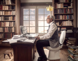 Ein älterer, schlanker und großer Mann sitzt vor einem vollen, unsortierten Schreibtisch in einem älteren Büro mit vielen vollen Bücherregalen, etwas unaufgeräumt. Der Mann: dunkle Hose und Schuhe, weißes Hemd, graue Weste, weiß-graue, nach hinten liegende Haare, Brille, Blick ernst und nachdenklich etwas nach oben in den Raum. Vor dem Schreibtisch ein kleiner Hocker, vielleicht als Möglichkeit, darauf zu knien. Über dem Kopf des Mannes schwebt mit etwas Abstand eine goldene Krone mit roten Edelsteinen. Der Schreibtischstuhl, auf dem der Mann sitzt, besteht aus einer klassischen Toilette.